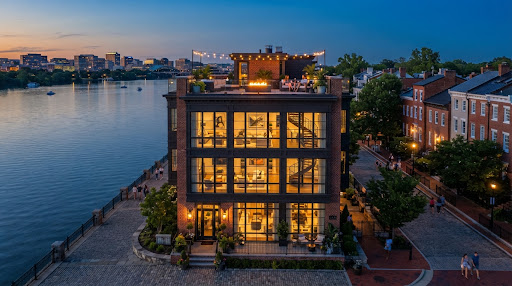 Luxury waterfront rowhouse in Old Town Alexandria Virginia overlooking the calm Potomac river at twilight, featuring rooftop deck with amber fire table and expansive floor to ceiling glass windows